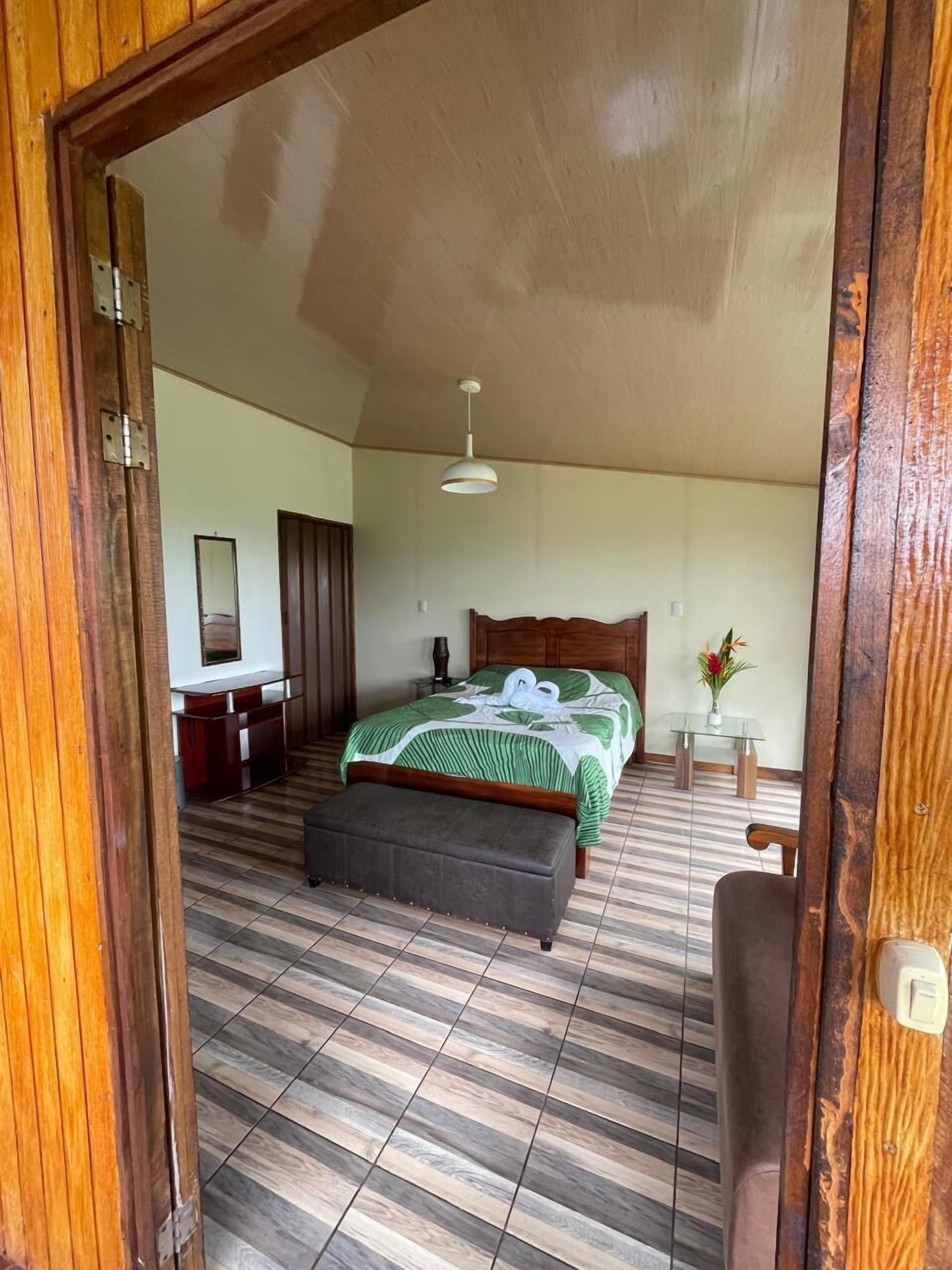 A bedroom with a bed, a bedside table, a mirror, a hanging light fixture, and a wooden door frame.