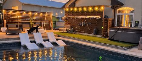 A swimming pool with lounge chairs, a covered patio area with string lights, and a building with a chimney.