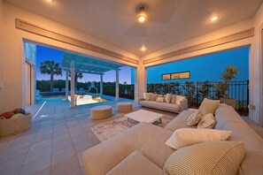 Un salon moderne avec un canapé, des poufs et une table basse. Une grande fenêtre offre une vue sur une piscine et des palmiers.