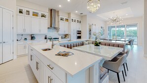 A modern kitchen with white cabinetry, a central island, and a dining area with a large window. - 9BR Reunion Mansion | Pool, Spa & Home Theater (Kissimmee)