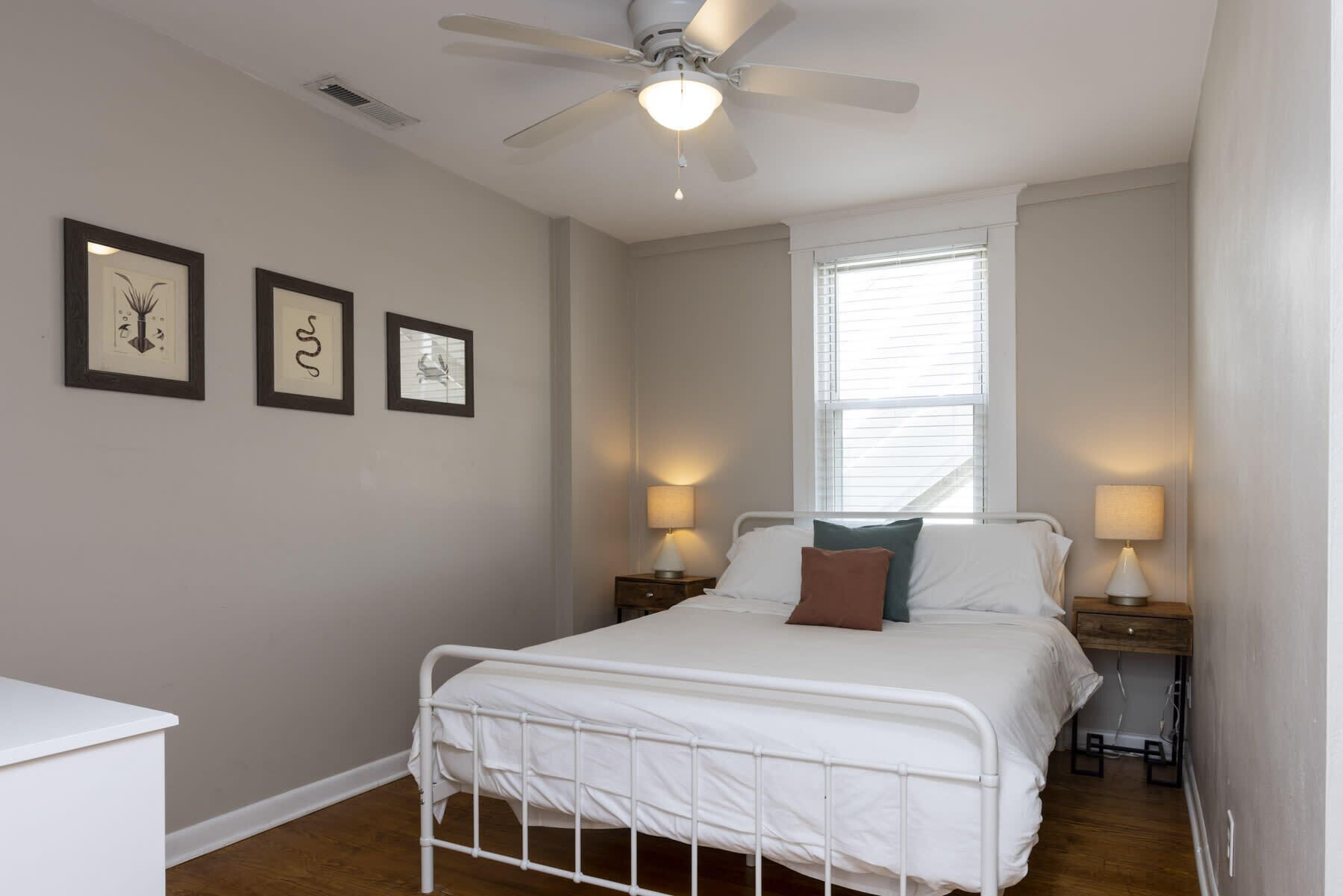 A bedroom with a bed, two bedside tables with lamps, a ceiling fan, and three framed pictures on the wall.