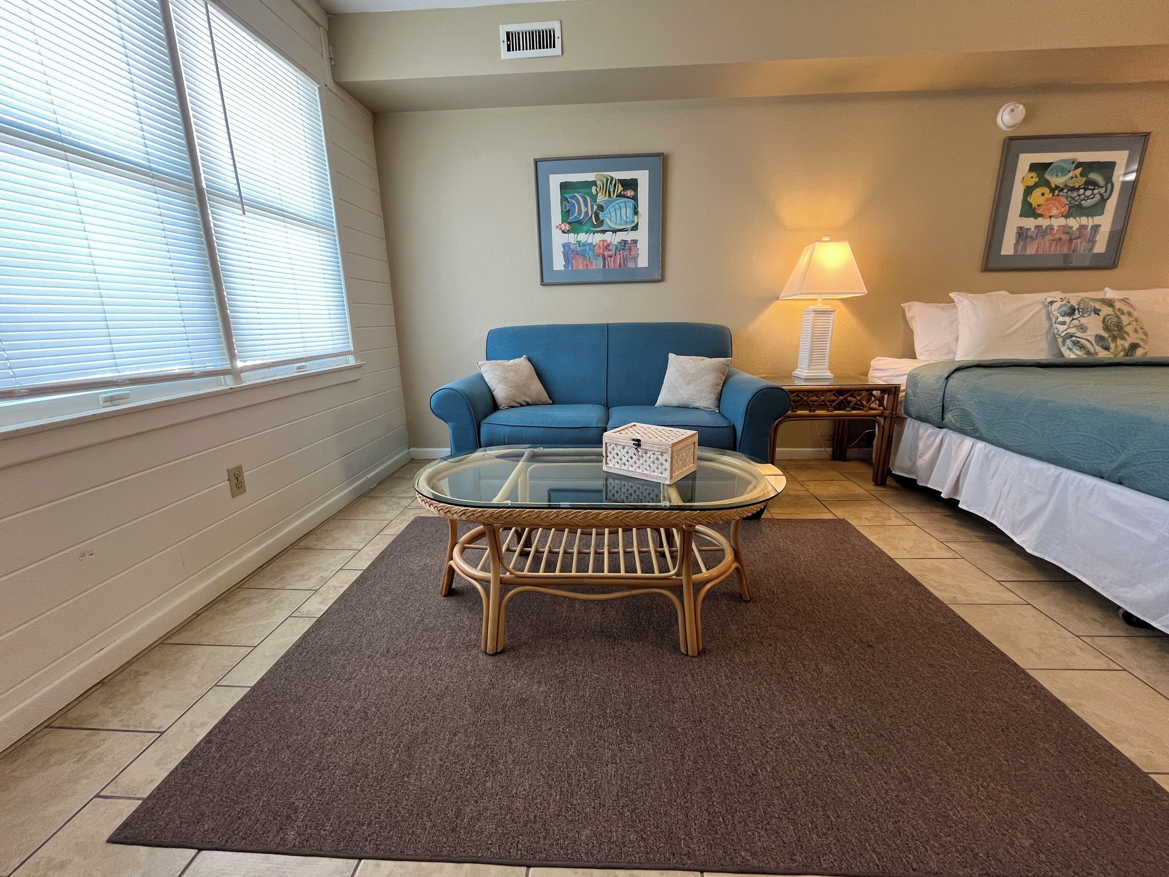A cozy room with a blue sofa, a glass coffee table, a bed with a green bedspread, and two wall art pieces.