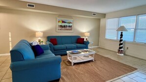 A living room with two blue sofas, a glass coffee table, and a rug. There is a painting on the wall and a lamp near the window. - Cape Hatteras Motel (Buxton)
