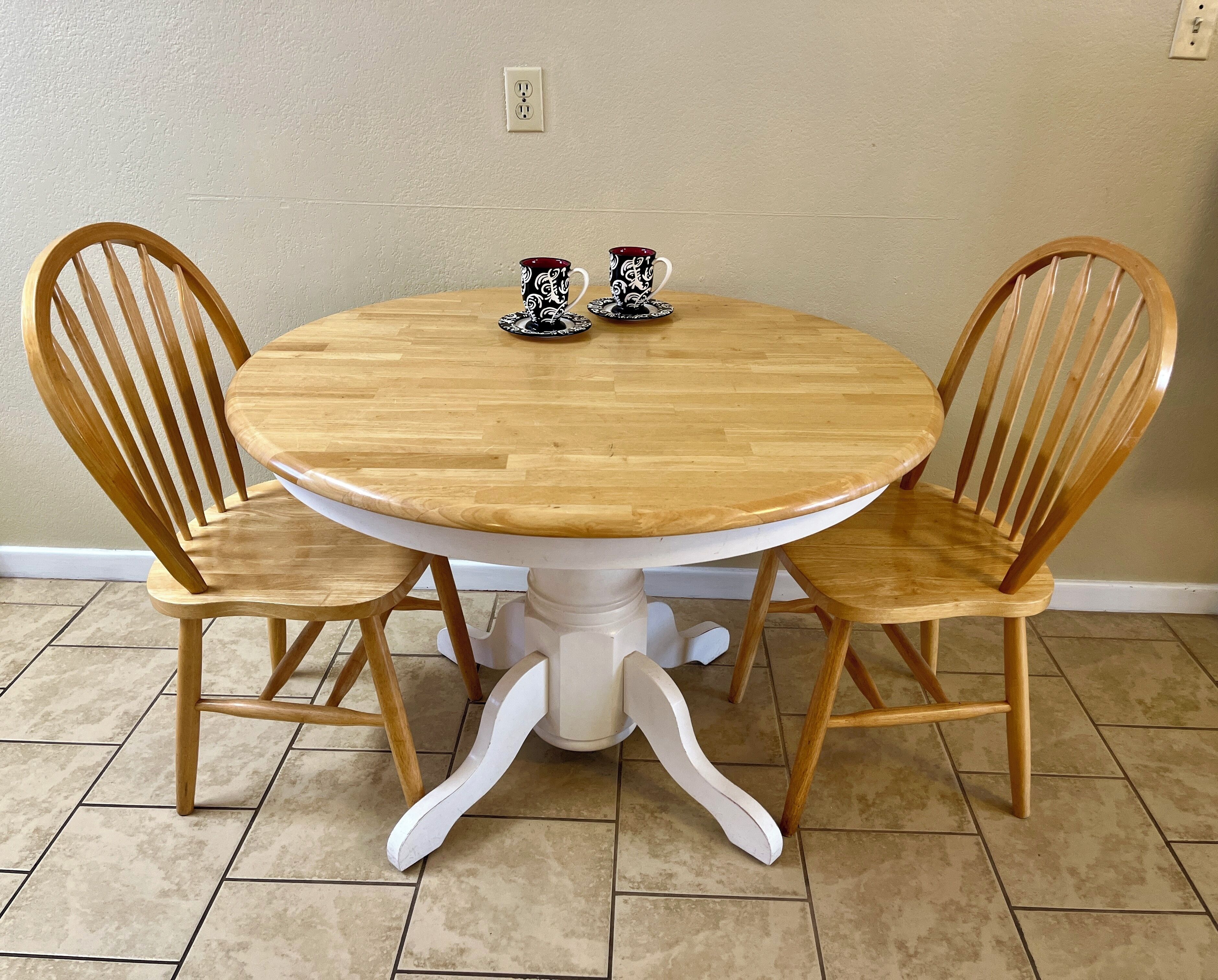 A round wooden table with two matching chairs, two patterned cups, and a white base.
