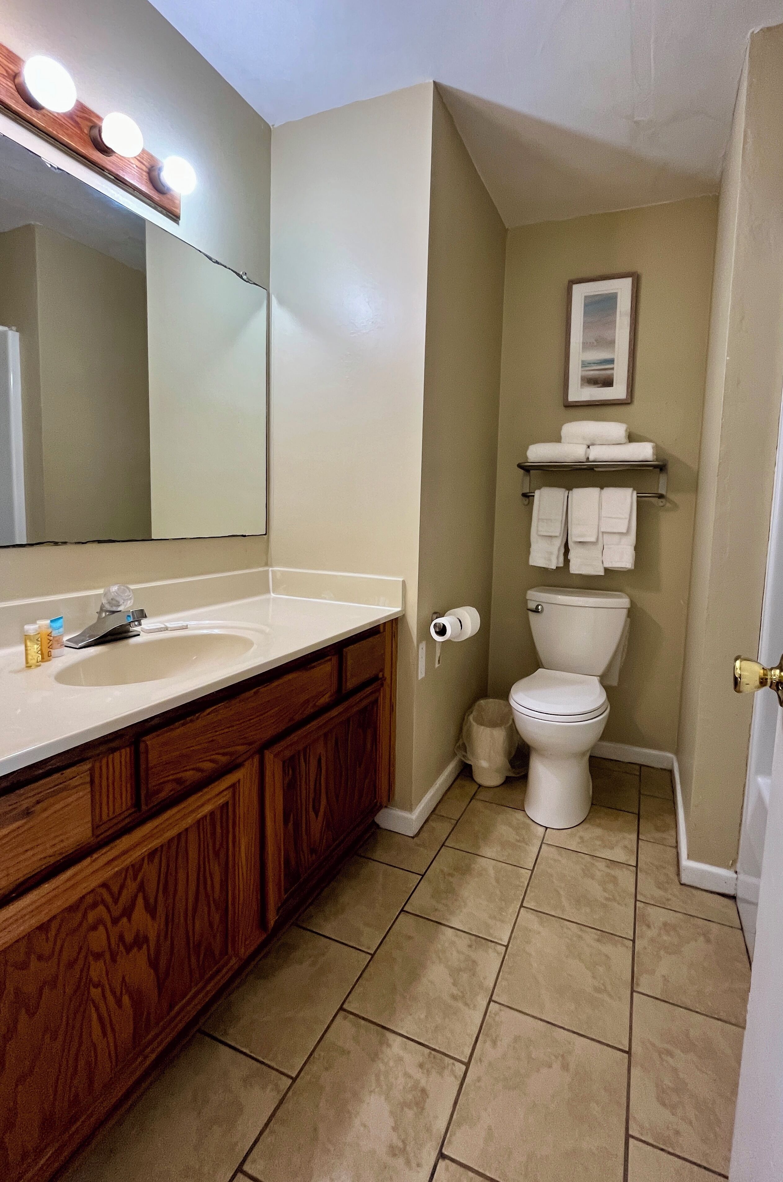 Un baño con tocador de madera, un lavabo, un espejo con luces, un inodoro y un estante con toallas.