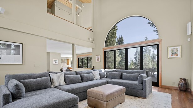 Ein geräumiger Wohnbereich mit einem großen Ecksofa, dazu passendem Hocker und einem großen Bogenfenster mit Blick auf Bäume.