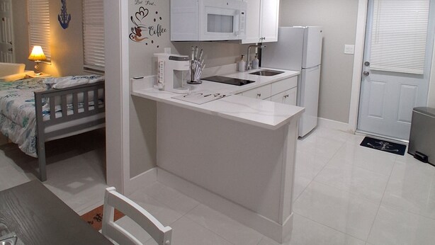 Een compacte keuken met witte kastjes, een magnetron en een koelkast. Er is een eethoek met een tafel en stoelen, en een bed in de aangrenzende kamer.
