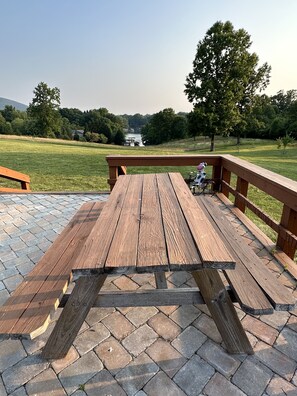 Een houten picknicktafel met een metalen leuning, geplaatst op een verhard gebied met uitzicht op een meer en bomen.