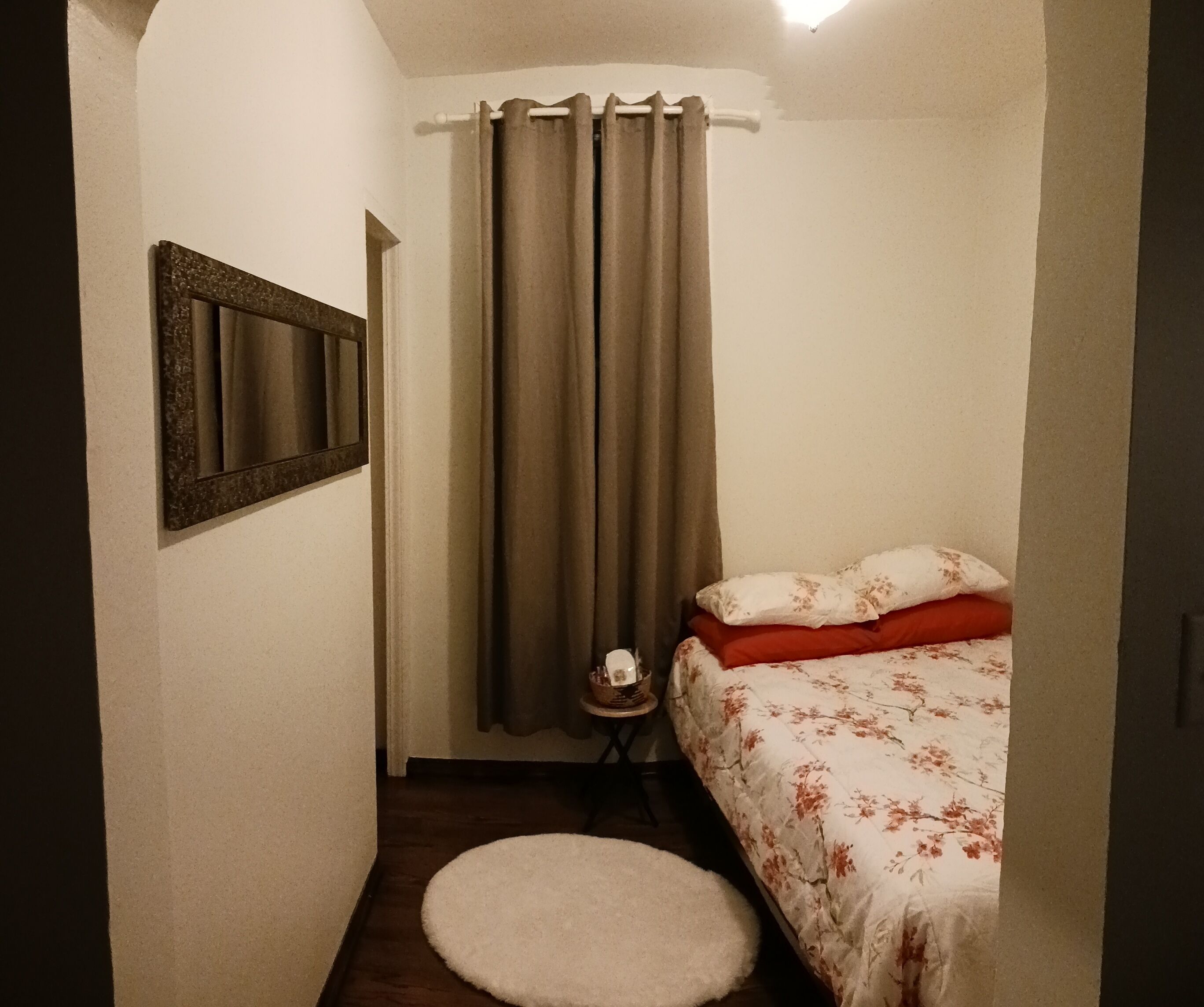 Une petite chambre avec un lit, un tapis rond, un miroir et un rideau.