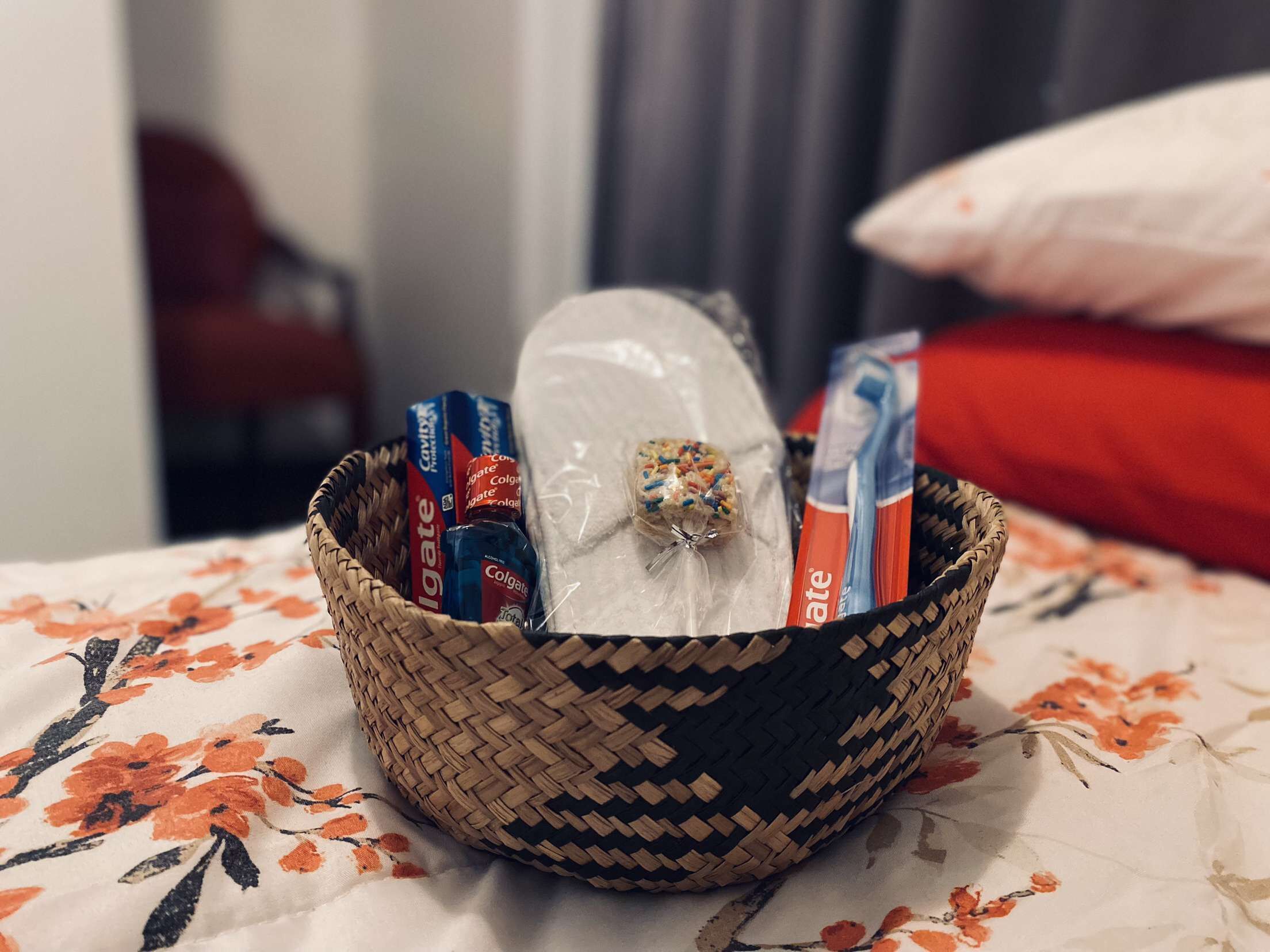 A woven basket on a bed with a floral bedspread, containing toiletries including toothpaste and a decorative item.