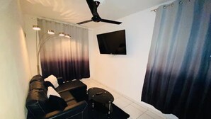 Un salon avec un canapé noir, un petit tabouret noir, un ventilateur de plafond, un téléviseur à écran plat fixé au mur et une fenêtre avec des rideaux.