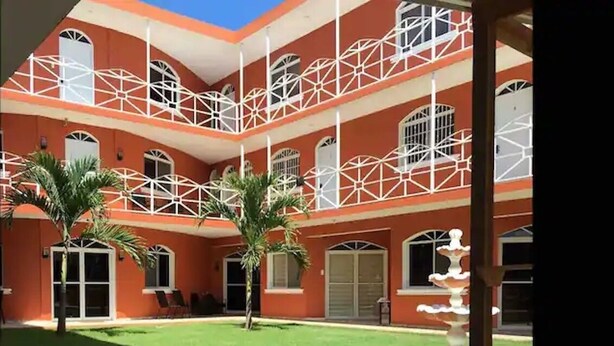 Un immeuble à plusieurs étages, doté d’une façade rouge, de balcons blancs et d’une cour intérieure agrémentée de palmiers et d’une fontaine.