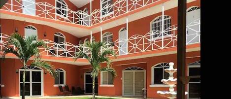 A multi-story building with a red facade, white balconies, and a central courtyard with palm trees and a fountain.