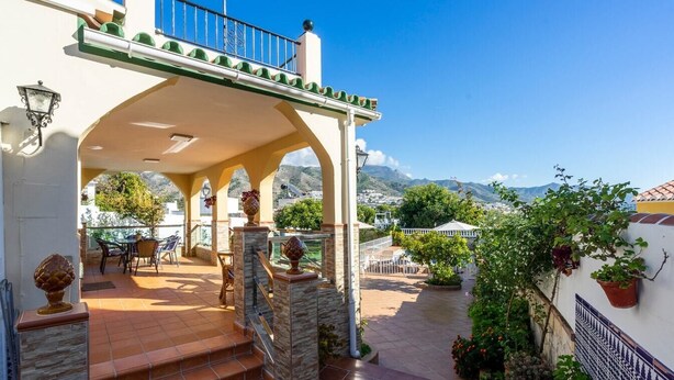 Un patio techado al aire libre con vistas a montañas, columnas de piedra y una mesa con sillas.