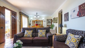 Una sala de estar con un sofá marrón, cojines estampados, una mesa de centro con un jarrón de flores y un comedor al fondo.