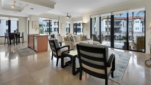 Un vaste espace de vie comprenant un canapé, des fauteuils et une table à manger. Il y a également un coin cuisine avec un comptoir et des placards. De grandes fenêtres offrent une vue sur l’extérieur.