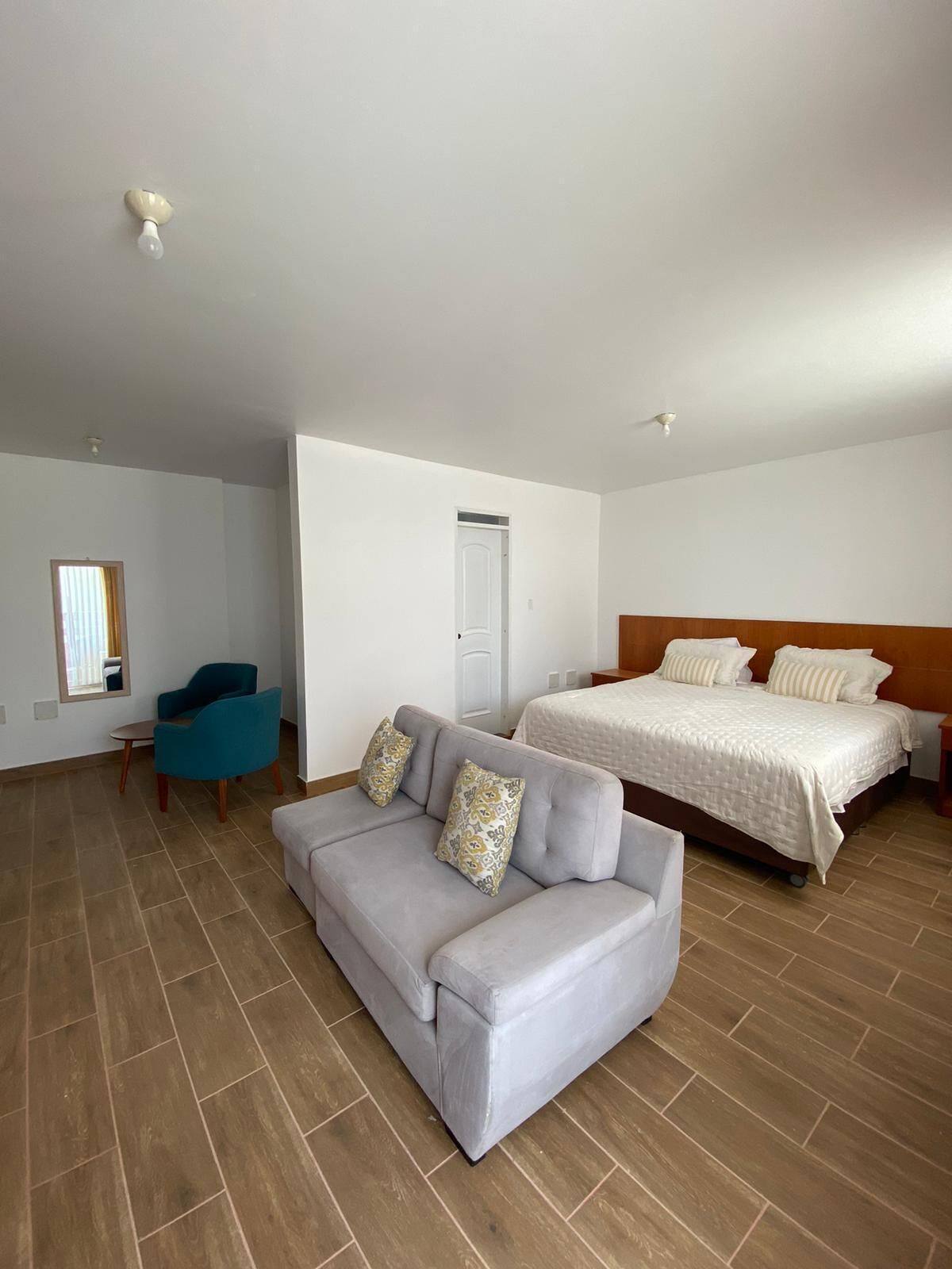 Una habitación con un sofá gris, una cama con ropa de cama blanca, una silla azul y suelo de madera.