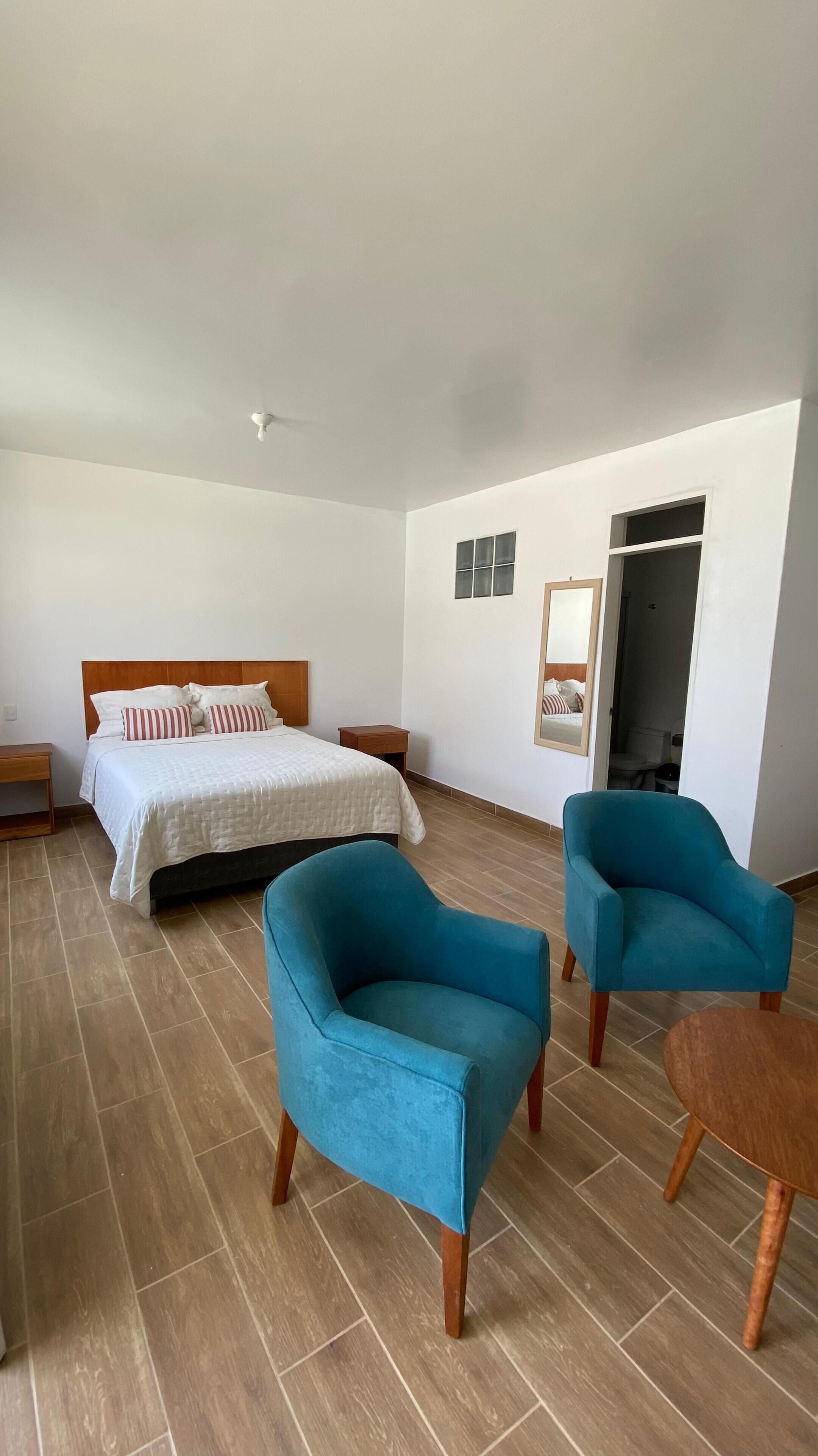 Habitación de hotel con una cama, dos sillones azules, una mesa de madera y un baño pequeño visible a través de una puerta abierta.