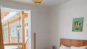 A bedroom with a large bed, wooden flooring, a ceiling light fixture, a painting on the wall, and a view of the outside through a window.
