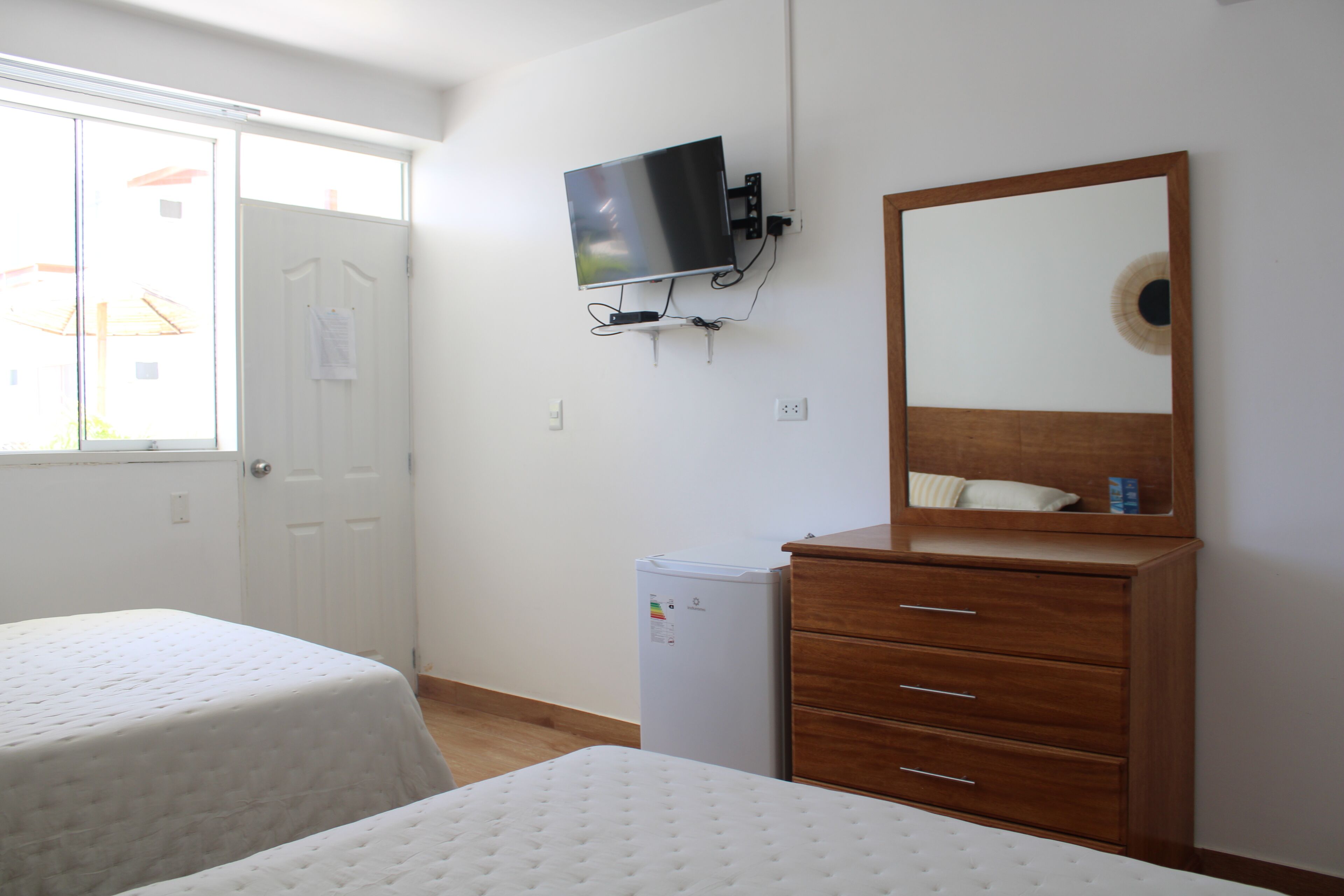 A bedroom with two beds, a TV mounted on the wall, a small refrigerator, and a wooden dresser with a mirror.