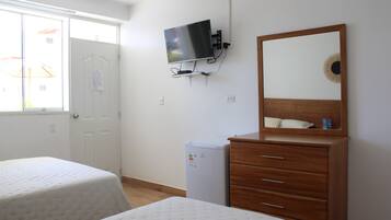 Quarto com duas camas, televisão fixada na parede, pequeno frigorífico e cómoda de madeira com espelho.