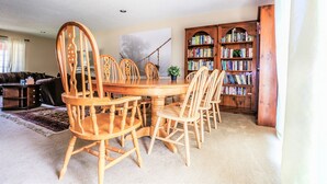 Een eetgedeelte met een ronde houten tafel en stoelen, een boekenkast en een bank op de achtergrond.