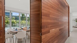 A modern dining area with a wooden partition, a round table with chairs, and a view of palm trees through large windows. - KARAT Villa Splendida (Playa Paraiso)
