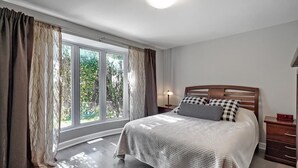 Un dormitorio con un ventanal grande, una cama de madera, una mesita de noche y un aparador.