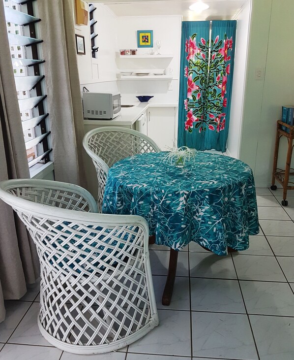 A dining area with a round table covered in a floral tablecloth, two wicker chairs, a microwave, a small shelf with dishes, and a decorative curtain.