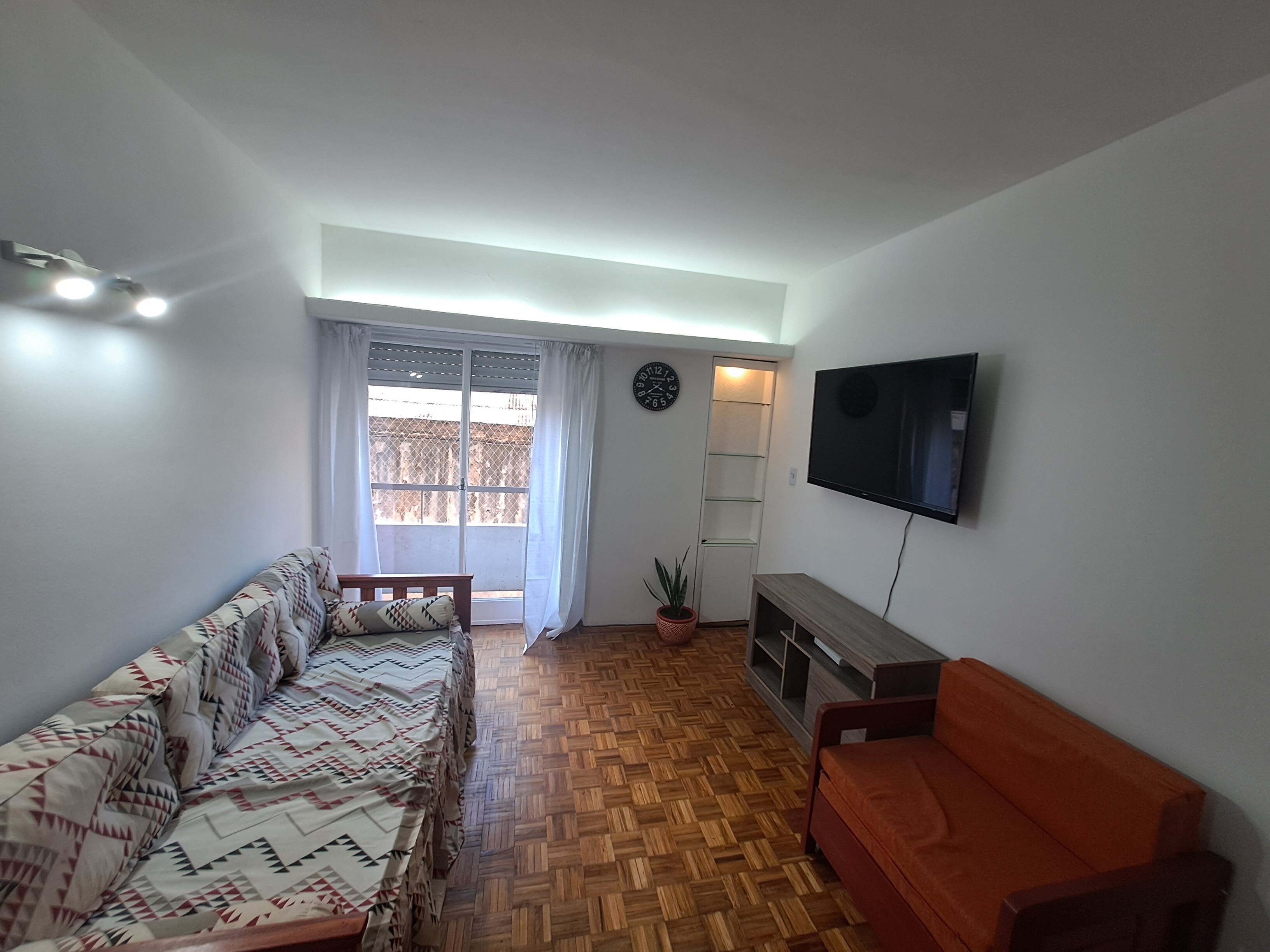 Una sala de estar con un sofá, un televisor, un banco de madera, una planta en maceta y un ventanal corredizo que da a un balcón.
