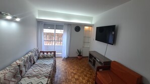 Een woonkamer met een bank, een televisie, een houten bankje, een potplant en een schuifdeur naar een balkon.