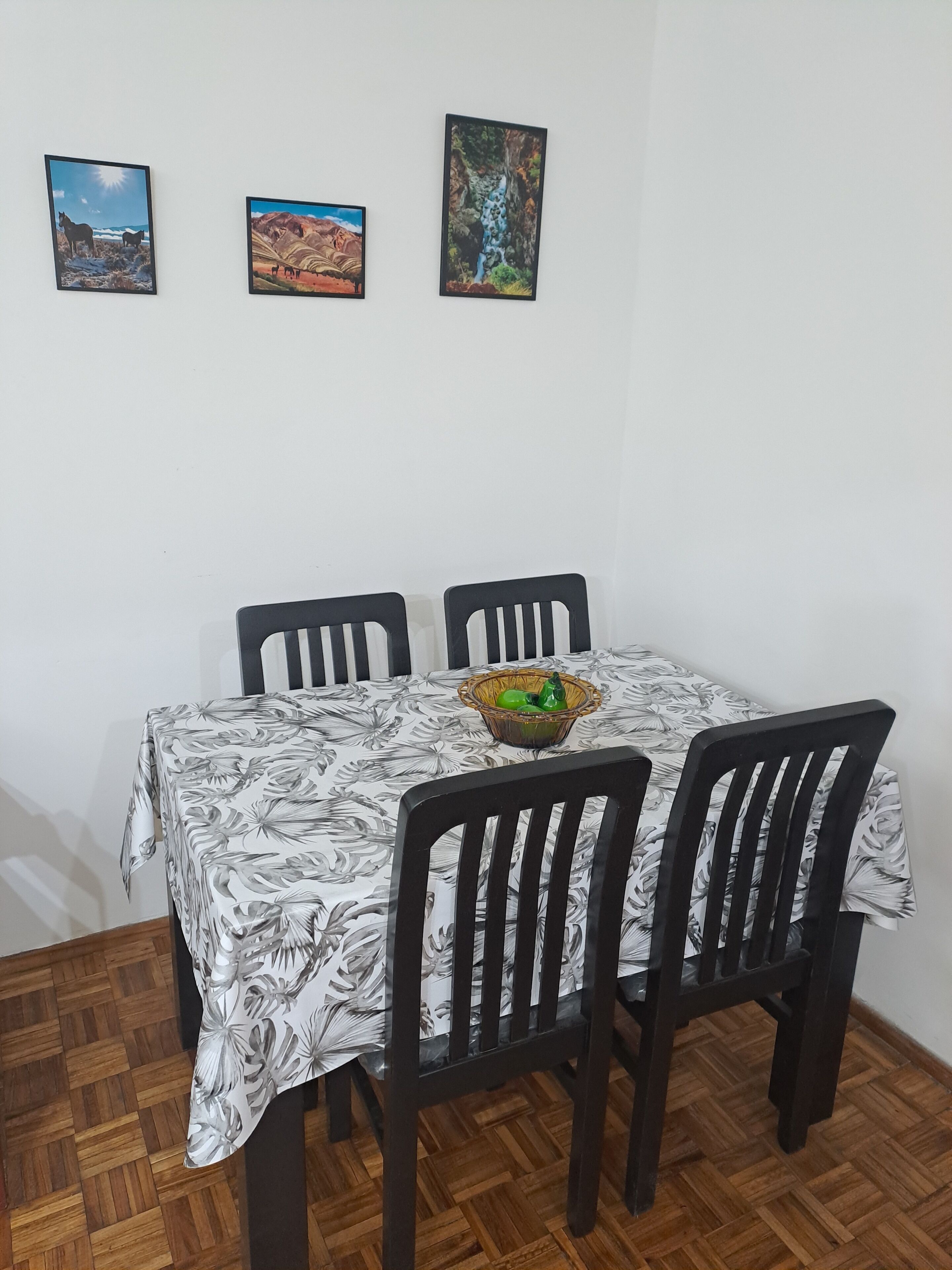Un comedor con una mesa cubierta por un mantel floral, rodeada por cuatro sillas. Hay tres cuadros en la pared sobre la mesa.