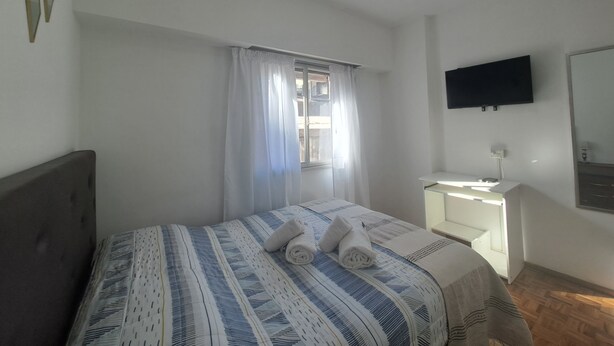 Een slaapkamer met een bed, nachtkastje, een aan de muur bevestigde televisie en een raam met gordijnen.