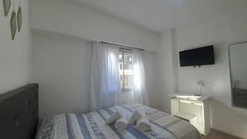 Un dormitorio con cama, mesita de noche, televisor montado en la pared y ventana con cortinas.