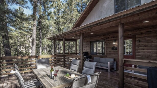 A wooden deck with a dining table and chairs, a sofa, and a window overlooking a forest. - Bearadise - Upper Canyon Gem, Hot Tub, Game Room, HUGE deck (Ruidoso)