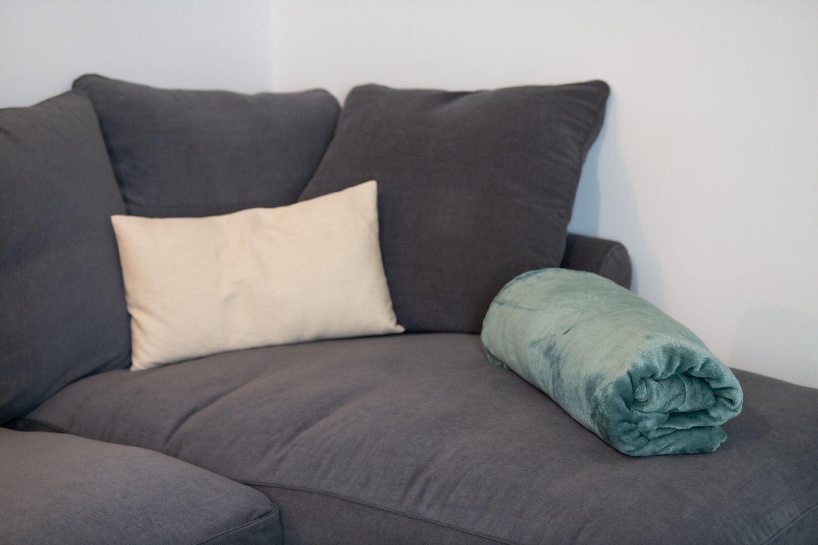 A grey sofa with a beige pillow and a rolled-up teal blanket.