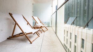 Un couloir avec deux fauteuils en bois et une petite table, qui mène à un mur de verre donnant sur un immeuble.