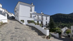 Un bâtiment blanc avec un escalier en pierre menant à l’entrée, entouré de verdure et avec une montagne en arrière-plan.