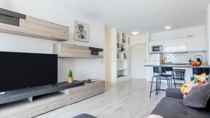 A modern living room with a flat-screen TV, a grey sofa, and a dining area with a kitchen in the background.