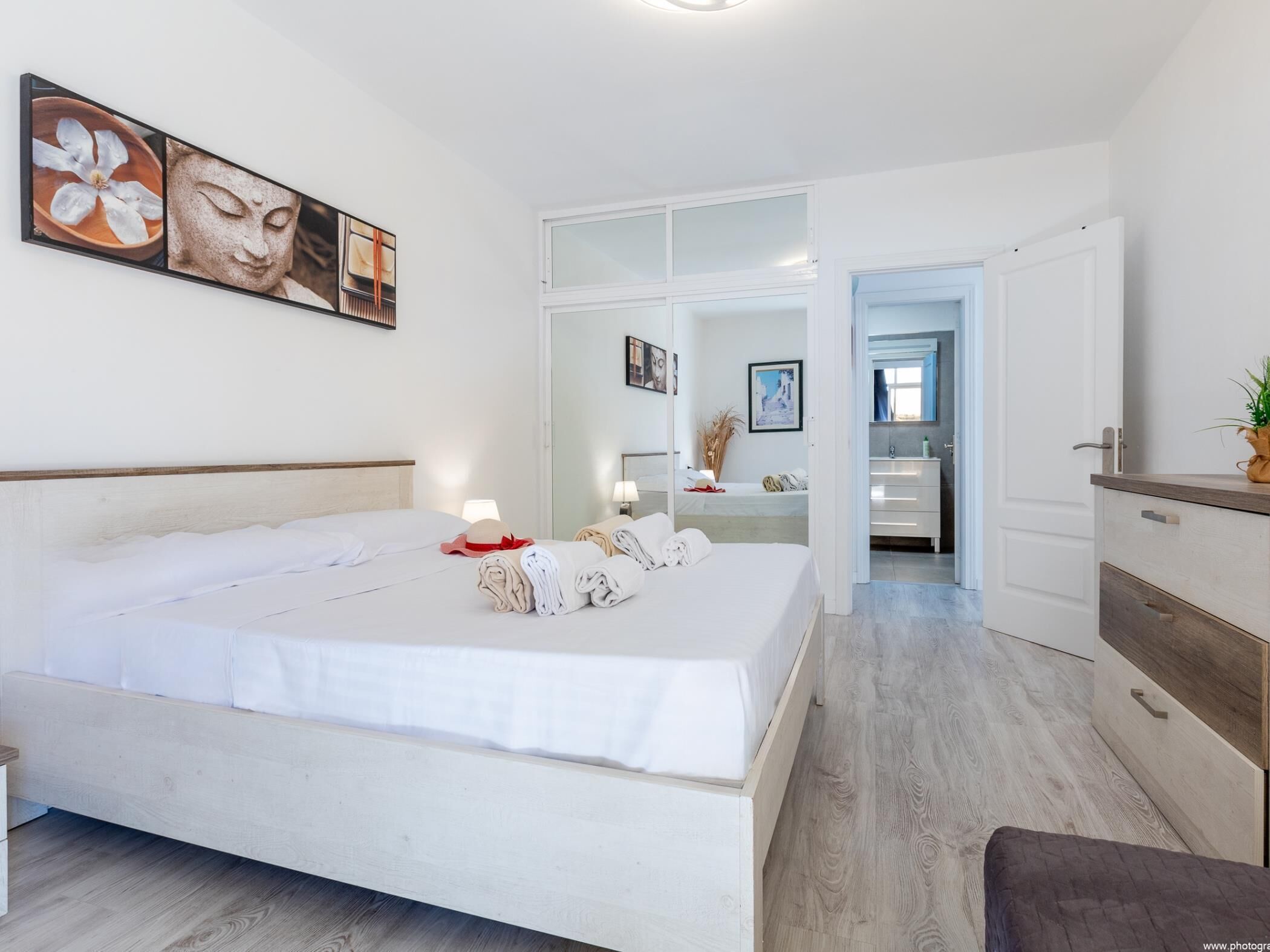Une chambre d’hôtel moderne avec un grand lit, une table de chevet avec une lampe, une salle de bain visible à travers une porte ouverte et des œuvres d’art accrochées aux murs.