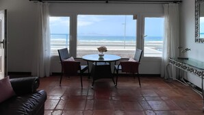 Ein Zimmer mit Blick auf den Strand, ein runder Tisch mit Stühlen, eine Vase mit Blumen und ein schwarzes Ledersofa.
