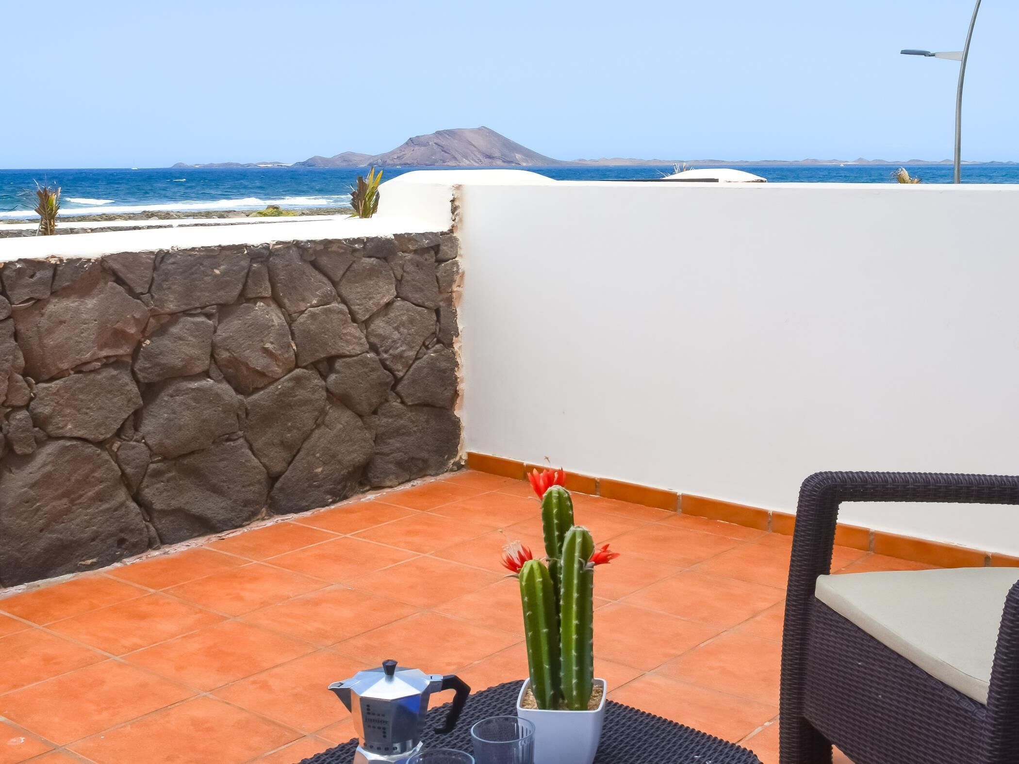 A balcony with a stone wall, a cactus, a coffee pot, and a wicker chair.