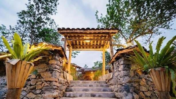 Una escalera de piedra conduce a una pérgola de madera con techo de tejas, rodeada de follaje.