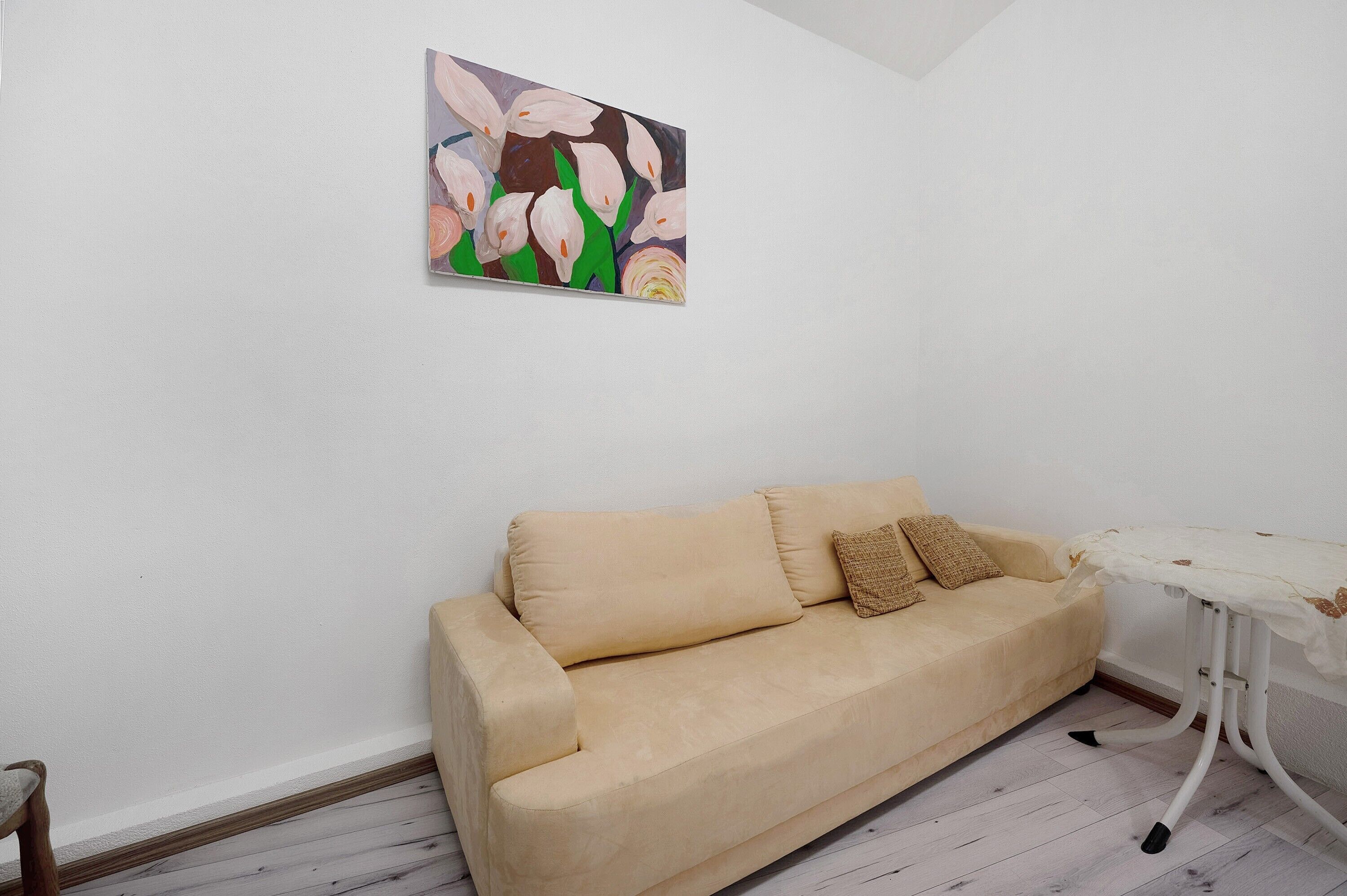 A beige sofa with a cushion, a small table with a white cloth, and a painting of flowers on the wall.