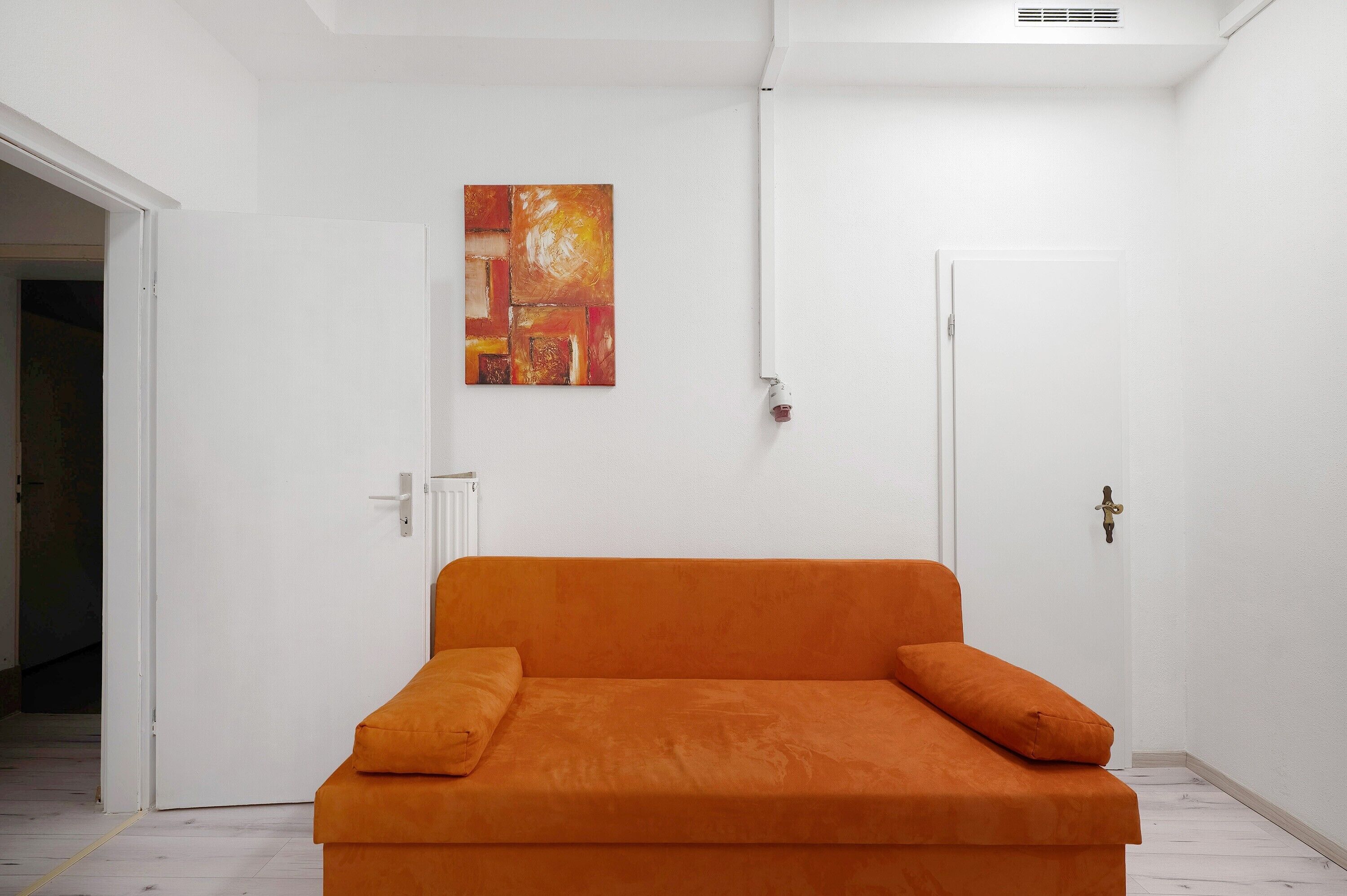 Ein Zimmer mit einer weißen Tür, einem Heizkörper und einem einzelnen orangefarbenen Sofa mit Kissen.