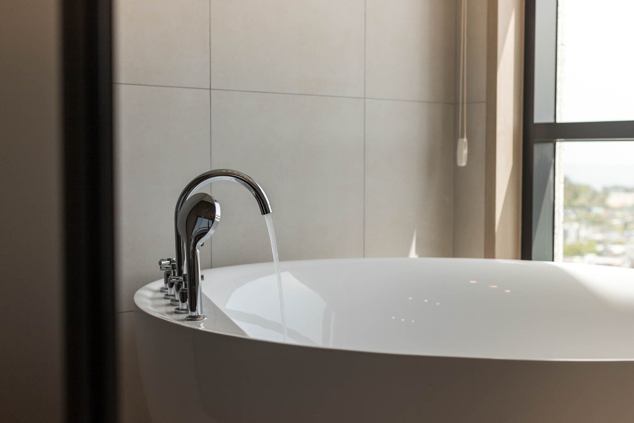 A modern bathroom with a large bathtub, a chrome faucet, and a window providing a view of the city.