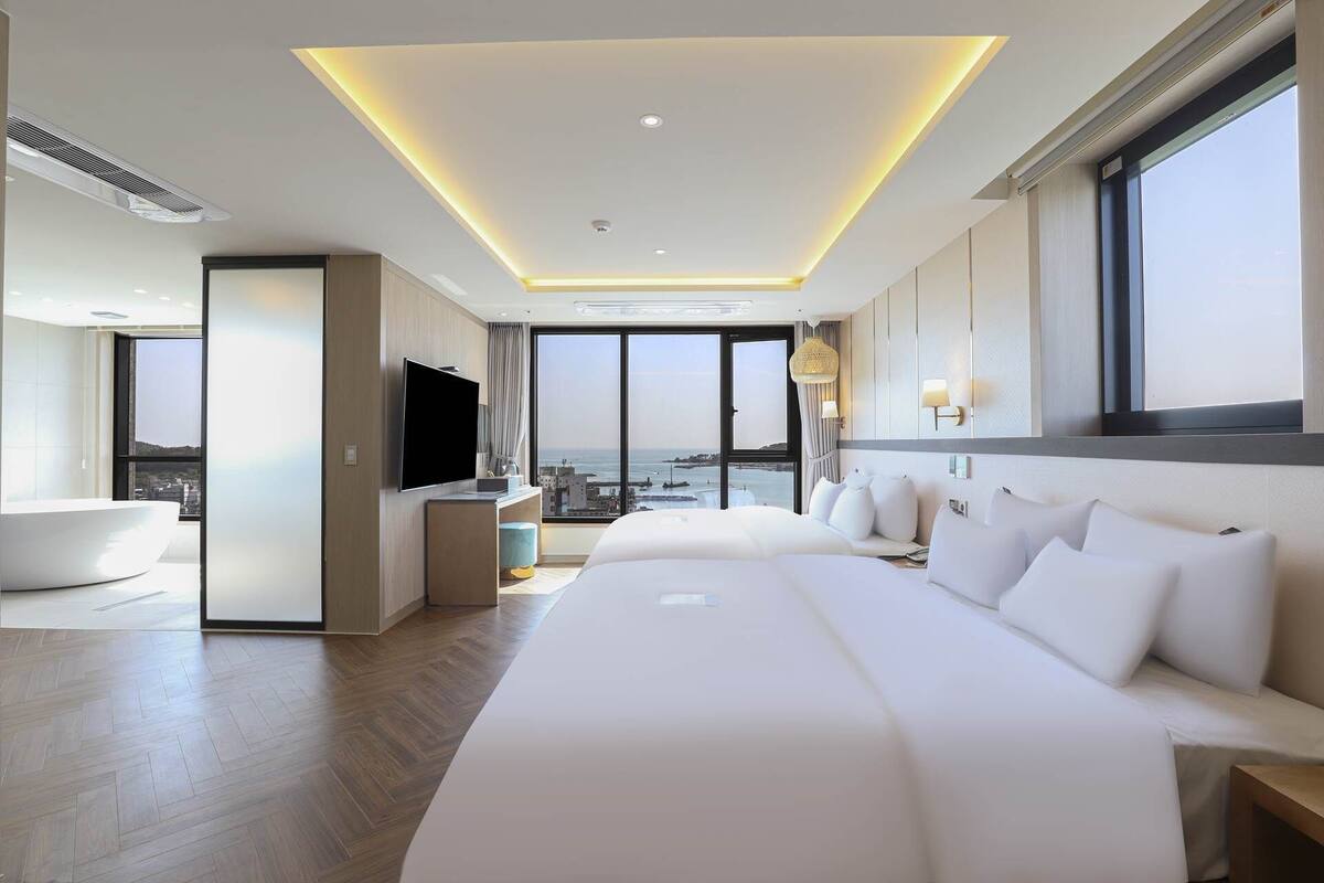 A modern hotel room with a large bed, a TV, a bathtub, and a city view.