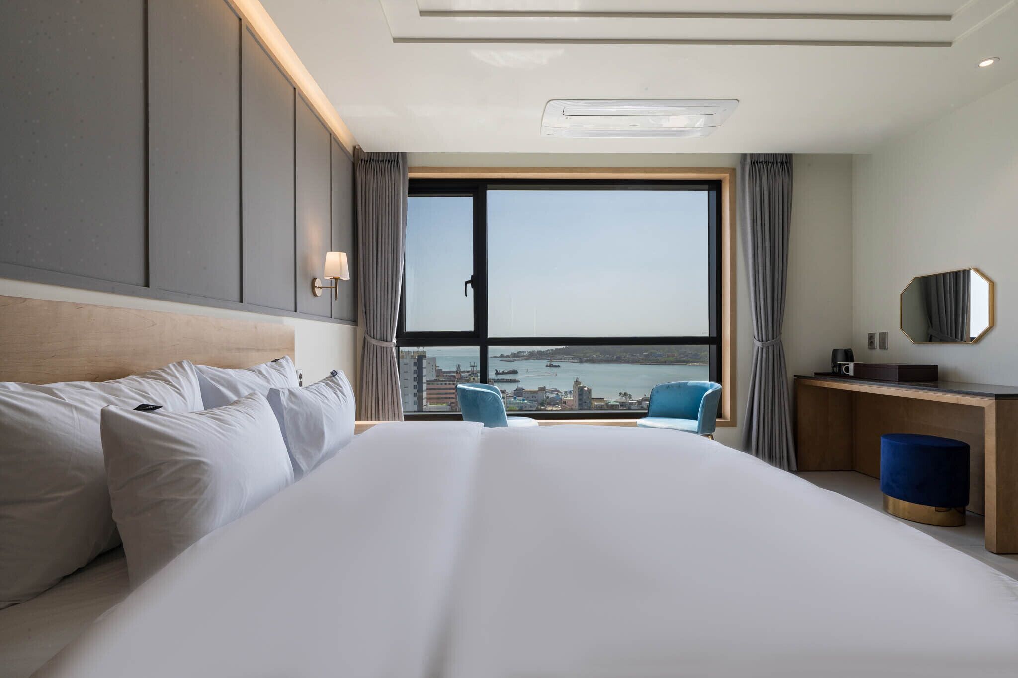 Habitación de hotel con una cama grande, un escritorio, una silla y vistas a un paisaje urbano a través de un ventanal amplio.