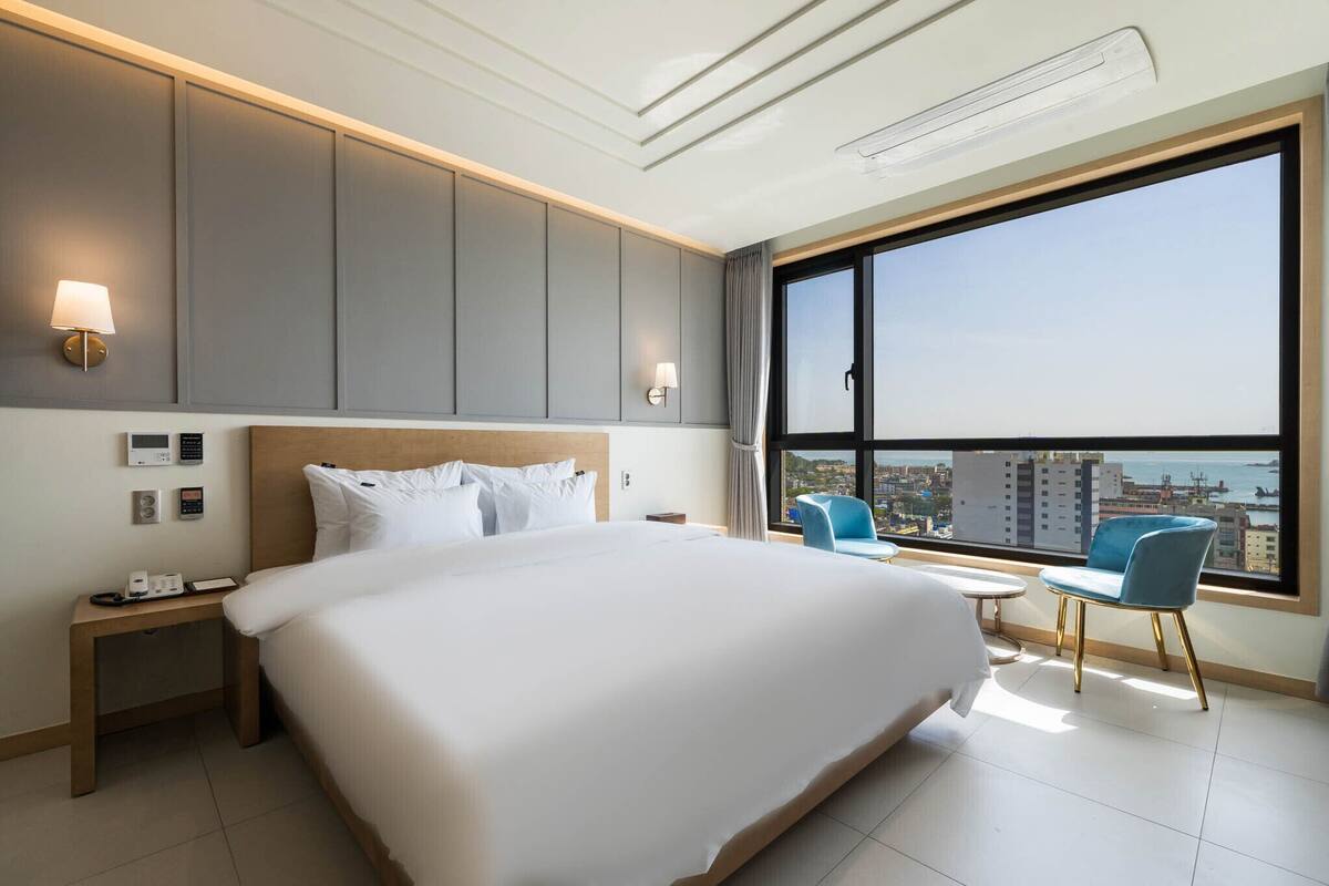 A hotel room with a large bed, bedside tables, a desk, and a view of the city through a large window.