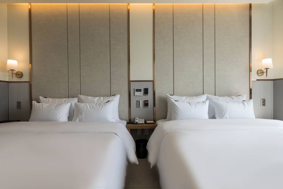 A hotel room with two beds, a control panel, and wall-mounted lamps.